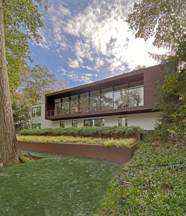 Exterior view of a contemporary two-storey residence set within a mature landscaped garden. The building has a clean rectangular form, a broad projecting upper-level volume clad in dark brown material, extensive floor-to-ceiling glazing with slim dark frames and a glass balustrade, and white rendered lower walls. Tiered rust-brown retaining planters, lawn, shrubs, and established trees surround the house.