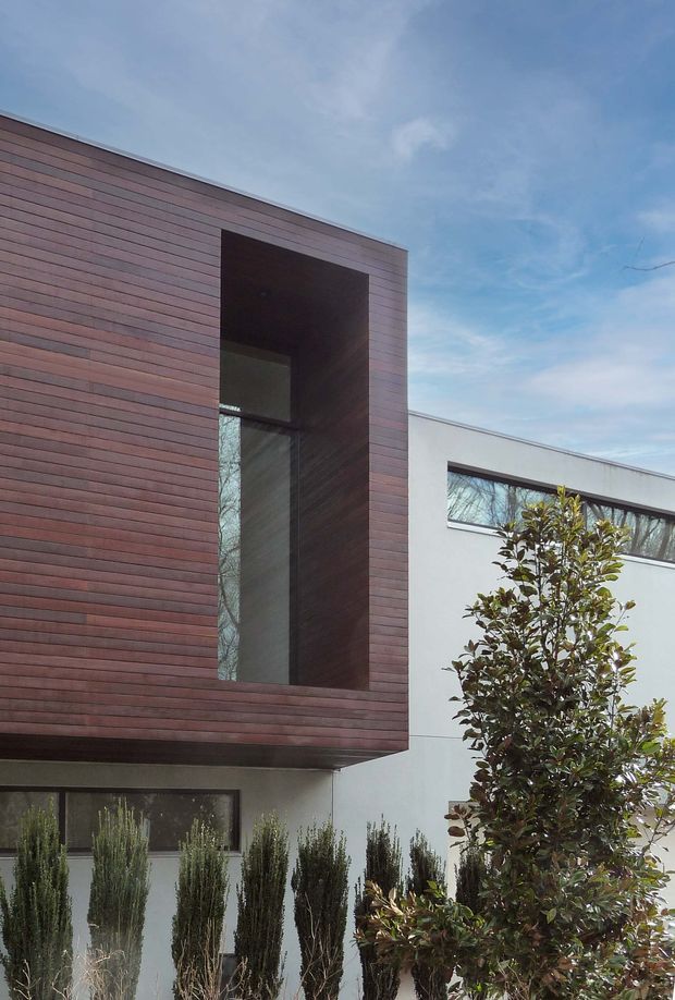 Exterior view of a contemporary multi-level residence featuring a projecting upper volume clad in horizontal dark timber boards, a deep recessed glazed opening, and adjacent smooth white rendered walls with narrow horizontal windows. Structured planting lines the frontage beneath a clear sky.