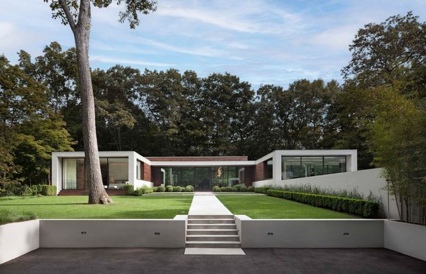 Front exterior view of a low, single-storey modern residence set within a landscaped suburban garden. The composition features a symmetrical arrangement of white rendered rectangular wings, extensive horizontal glazing, dark recessed central entry, flat rooflines, a straight paved path, clipped hedges, manicured lawn, and broad concrete retaining walls with steps leading up from the driveway.