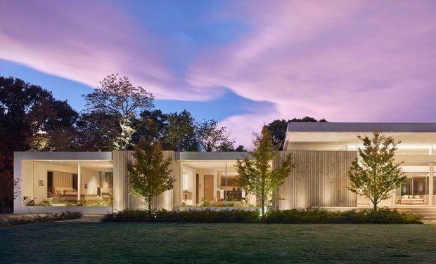 Dusk exterior view of a low-profile contemporary residence set behind a landscaped lawn. The house features broad floor-to-ceiling glazing, pale vertical timber-like wall cladding, deep roof overhangs, and connected single-storey volumes with warm interior lighting visible through the windows. Mature trees and low planting frame the facade.