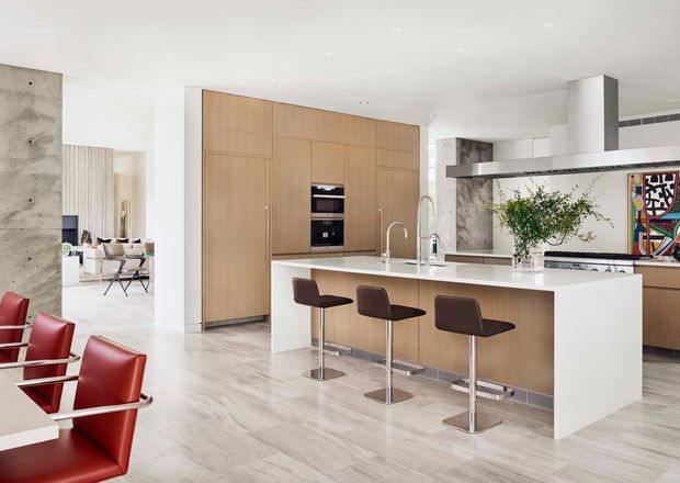 Bright, spacious contemporary kitchen with a large central island and three bar stools. The room features full-height light timber cabinetry, a white waterfall-edge island, pale stone-look flooring, a linear stainless-steel rangehood, integrated appliances, and a light neutral palette. Red dining chairs and colourful wall art provide accents, while an open dining area is visible beyond the kitchen.