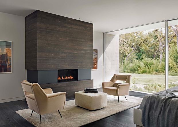 A refined contemporary living room with two upholstered lounge chairs and a large ottoman arranged around a linear fireplace. The fireplace is integrated into a dark timber-toned feature wall and flanked by dark cabinetry or wall panels. Floor-to-ceiling glazing opens the room to a leafy garden, while pale walls, a neutral rug, and dark flooring create a restrained, sophisticated interior.