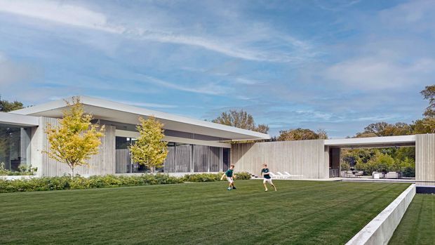 Wide exterior view of a low, single-storey contemporary residence with long horizontal roof planes, extensive glazing, pale vertical cladding and a covered carport or outdoor pavilion at right. A manicured lawn dominates the foreground, with young ornamental trees and planted borders around the building.