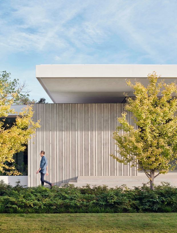 A contemporary low-profile building exterior viewed from a landscaped lawn. The facade combines a broad flat projecting roof with a deep soffit, pale smooth rendered surfaces, and a tall vertically ribbed concrete or textured wall. Dark recessed glazing is partly visible behind mature trees and planting. The composition is clean and minimalist, with a pedestrian providing scale.