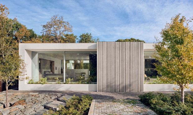 Single-storey contemporary residential facade set within landscaped grounds. The flat-roofed white structure has broad floor-to-ceiling glazing on either side of a prominent vertically ribbed grey feature screen, with a paved approach, stone landscaping and mature trees in the foreground. Interior furnishings are faintly visible through the glazing.