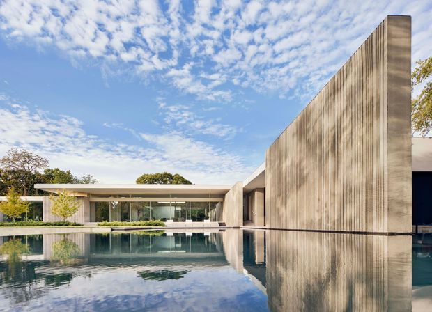 A striking low-slung modern residence arranged around a reflective pool. The composition features a long glazed pavilion, broad flat rooflines, and a tall vertically ribbed concrete or rendered wall forming a strong sculptural edge. The restrained neutral architecture is mirrored in calm water, with landscaped planting and a bright blue sky in the background.