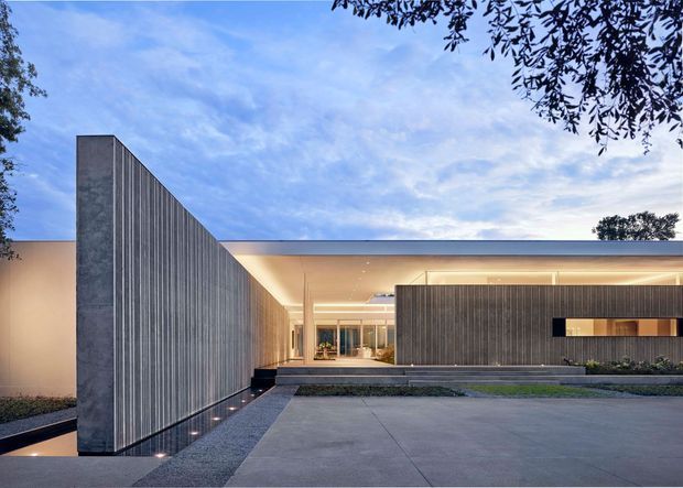 A contemporary single-storey residence viewed from the front, with a long low flat roof, expansive recessed entry glazing, and dramatic vertical dark timber or metal screening walls. Broad concrete paving and carefully integrated linear lighting emphasize the minimalist architectural composition, while mature trees frame the site.