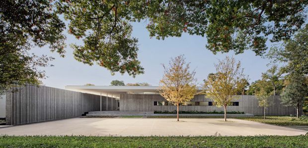 A wide exterior view of a low, modern single-storey residence set behind a broad paved forecourt. The building has a long, shallow roof plane, extensive vertical grey cladding or screening, recessed glazing, and a sheltered entry/carport-like opening. Mature trees frame the site, with young deciduous trees planted along the front.