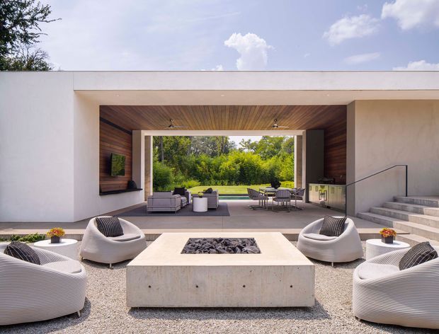 Contemporary indoor-outdoor living space opening onto a lush garden, with a large concrete fire table surrounded by curved woven lounge chairs and sofas. The covered entertaining area features warm timber-lined soffits and walls, white rendered walls, expansive glazing, an outdoor dining setting, and broad concrete steps leading upward.