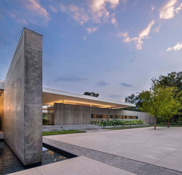 Exterior architectural photograph of a low, minimalist contemporary building at dusk. The composition features a tall monolithic concrete wall beside a shallow reflecting pool, a broad flat roof with concealed warm lighting, dark vertical timber cladding, extensive glazing, landscaped planting, and a large pale paved forecourt.