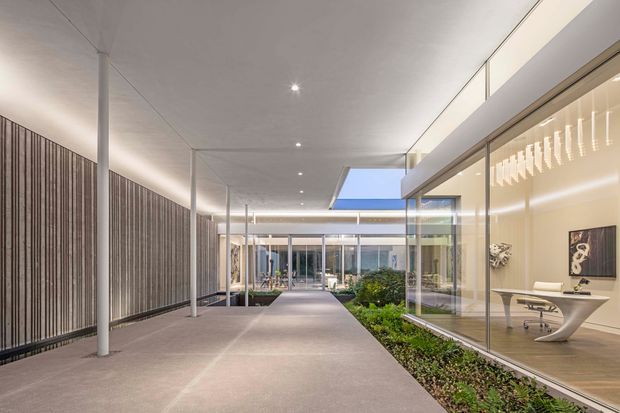 A refined contemporary interior courtyard or covered walkway with a long polished concrete floor, full-height glass walls, slim white structural columns, and a slatted timber feature wall. Landscaped planting runs through the central and edge areas, while a minimalist office or commercial interior with sculptural furniture is visible through the glazing. Recessed lighting and a narrow skylight create a bright, calm architectural setting.