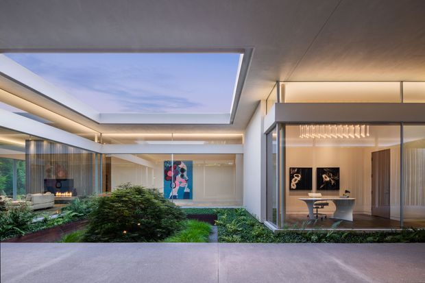 Contemporary modern residence organized around a landscaped internal courtyard. The view shows a broad open skylight, clean horizontal roof planes, extensive floor-to-ceiling glazing, warm concealed lighting, and a refined interior workspace visible through glass. Lush planting and a linear water feature soften the concrete and glass composition.