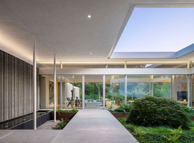Contemporary glazed courtyard passage with a broad concrete walkway leading through a landscaped internal garden. The space has full-height glass walls and sliding doors, slender metal posts, a large ceiling skylight, recessed lighting, and a dark vertical slatted feature wall. Dining furniture and a fireplace are visible beyond the glazing, with abundant greenery forming the central landscape.