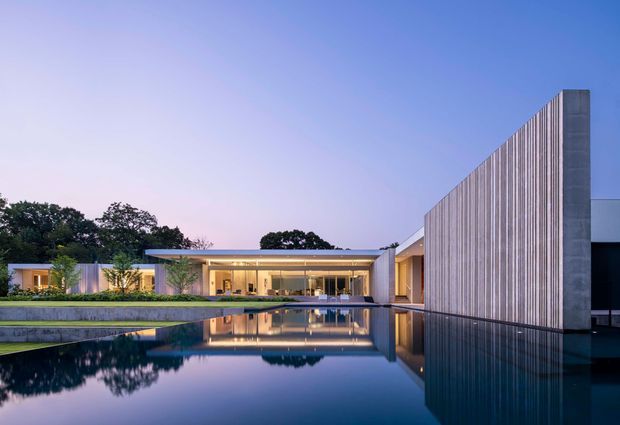 Twilight exterior view of a large contemporary single-storey residence, composed of low horizontal glazed volumes and a tall vertical screened wall. A long reflecting pool dominates the foreground, mirroring the architecture and sky, with landscaped grounds and mature trees behind the house.