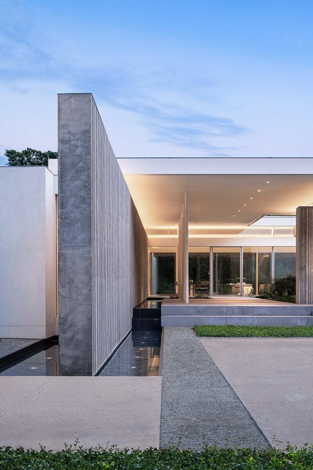 A refined contemporary residential exterior at dusk, featuring a strong vertical ribbed concrete privacy wall, smooth pale rendered volumes, a flat roof, broad recessed outdoor living area, extensive floor-to-ceiling glazing, and linear integrated ceiling lighting. Reflective dark water channels and geometric pale paving create a minimal courtyard approach with carefully clipped planting.