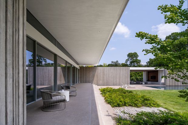 A contemporary single-storey courtyard residence viewed from a covered outdoor walkway. The image shows a broad concrete roof overhang, dark-framed floor-to-ceiling glazing, vertical timber-clad walls, a paved terrace with outdoor lounge chairs, and a landscaped lawn courtyard leading to a sheltered pavilion with a fireplace or dark feature wall. The composition emphasizes strong horizontal lines, indoor-outdoor connection, and restrained modern materials.