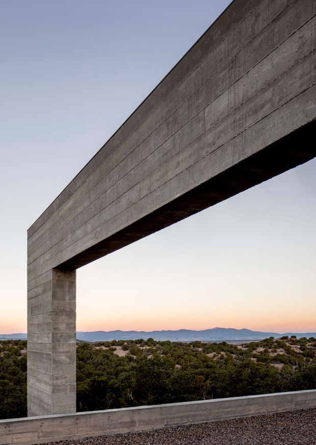 A minimalist exterior architectural detail featuring a monumental L-shaped concrete frame or portal opening toward a wide desert landscape. The exposed poured concrete has horizontal formwork lines and a raw, weathered grey finish. Low scrubland and distant mountains are visible through the opening beneath a pale sky, creating a strong geometric composition.