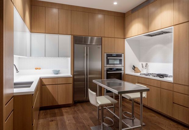A compact, highly organised contemporary kitchen with full-height warm timber cabinetry, integrated stainless-steel refrigerator and ovens, white upper cabinetry, and white textured tiled splashbacks. A central stainless-steel island with two stools provides preparation and dining space. Dark timber flooring, recessed ceiling lighting, and clean-lined built-in appliances create a warm, refined interior.