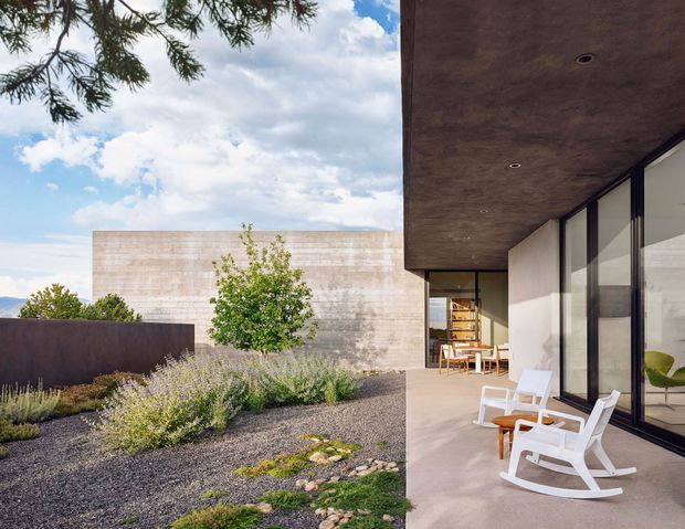 A contemporary low-profile residence with a broad cantilevered roof or soffit, floor-to-ceiling dark-framed glazing, and a minimalist courtyard terrace. The foreground features gravel landscaping with sculptural shrubs, grasses, rocks, and a small tree, while white outdoor rocking chairs and a timber table sit beneath the covered terrace. A pale textured masonry volume forms the backdrop, with distant landscape visible beyond the boundary wall.