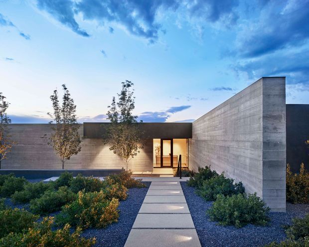 A minimalist modern single-storey residence viewed from the landscaped front approach at dusk. Strong horizontal and angular concrete walls frame a recessed, warmly lit entry with glazed timber-framed doors. A broad paved stepping-stone path crosses a gravel garden planted with low shrubs and slender trees, while the dramatic blue sky forms the background.