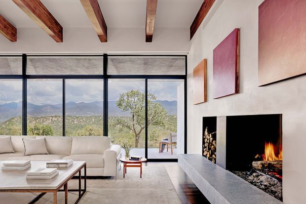 A bright contemporary living room with expansive black-framed floor-to-ceiling glazing overlooking a mountainous landscape. The room features a large pale sectional sofa, low coffee table, neutral rug, exposed timber ceiling beams, and a dramatic built-in black fireplace with a long grey stone hearth. Colourful abstract artwork adds accents to the white walls.