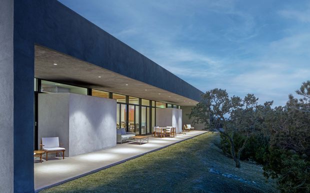 A low, minimalist modern residence viewed from the landscaped exterior. The dark blue-grey flat-roofed volume projects over a broad covered terrace with extensive floor-to-ceiling glazing, concrete or stucco walls, and restrained outdoor dining and lounge furniture. The building is set among mature shrubs and trees beneath an open sky, with warm interior lighting visible through the glass.