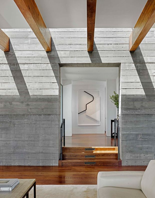 Interior view of a refined modern home showing a stepped transition between living and circulation spaces. Horizontal board-formed concrete walls frame a timber stair and raised landing, while substantial exposed timber ceiling beams cast strong shadows across the concrete. Warm timber flooring and stair treads contrast with white plaster surfaces, a pale abstract artwork, and partial contemporary seating in the foreground.