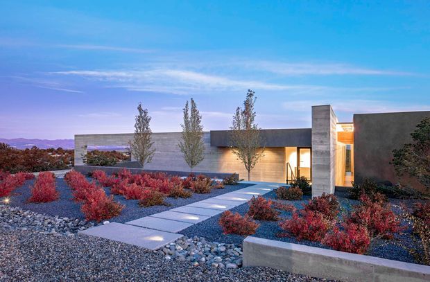 A low-slung modern residence viewed from the landscaped front approach at dusk. The house features flat roof forms, dark rendered walls, pale horizontal masonry or concrete walls, a tall projecting rectangular volume, and warm interior lighting visible through glazing. A straight paved walkway crosses a minimalist gravel garden with sculptural red shrubs, ornamental trees, rocks, and low concrete edging. Distant hills and a blue-purple sky form the background.