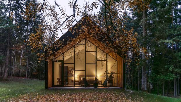 A small, contemporary gable-roof cabin or pavilion set within a wooded landscape. The dark timber-framed facade is almost entirely glazed, revealing a warmly lit minimalist living space with a sofa, lounge chair, shelving and plants. Fallen autumn leaves and dense trees surround the structure, creating a secluded woodland setting.