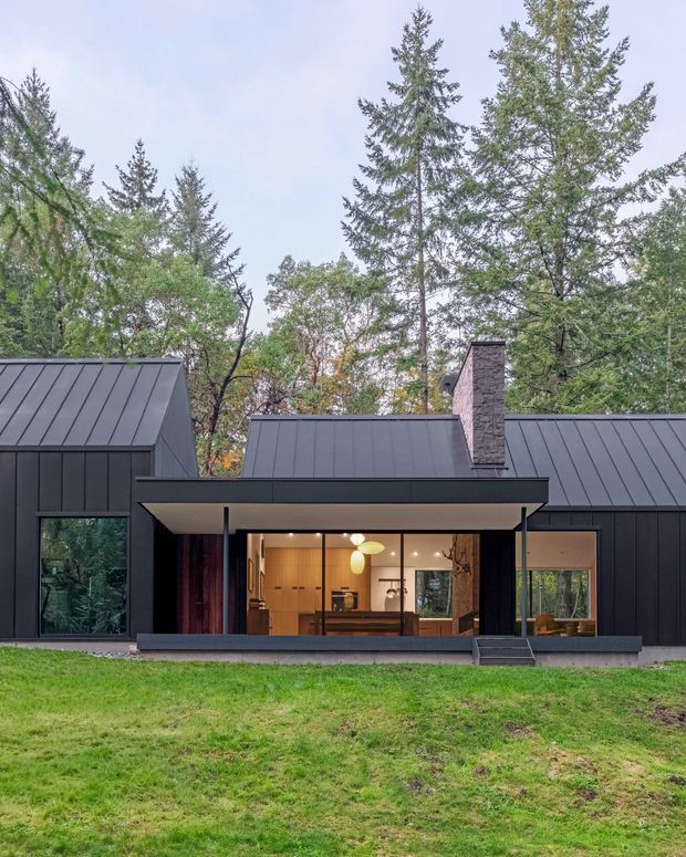 Front exterior view of a contemporary dark-clad house set within a mature wooded landscape. The single-storey composition has steep metal-clad roof forms, large glazed openings, a projecting covered entry or terrace, and a prominent stone-clad chimney. Warm interior lighting and timber finishes are visible through the central glazing.