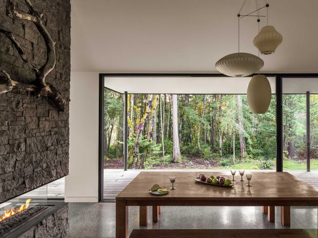 A refined dining area with a large solid timber dining table positioned beside a dramatic stone-clad fireplace. Full-height black-framed glazing opens the room visually toward a dense forest, while sculptural pendant lights hang above the table. The polished concrete or terrazzo-like floor, dark stone feature wall, warm timber, and neutral walls create a contemporary natural interior with strong indoor-outdoor connection.