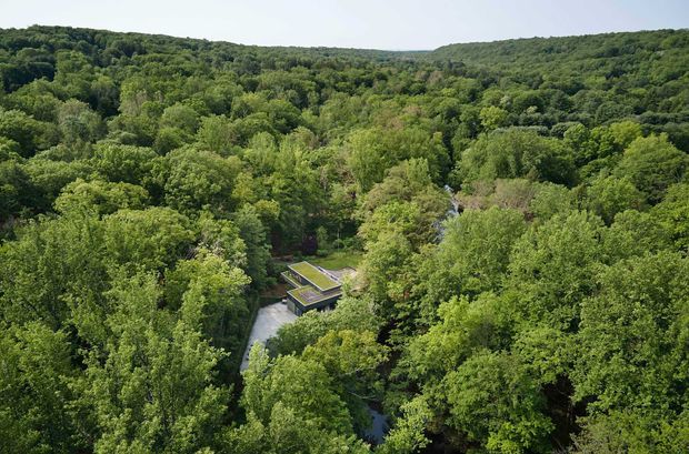 Aerial view of a modern low-profile building partially concealed within dense green woodland. The structure has flat roof planes, including a planted or green roof section, and is accessed by a narrow driveway or path. Rolling forested hills extend across the background.