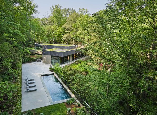 Elevated view of a modern, flat-roofed house nestled within dense mature woodland. The dark contemporary building has extensive glazing and projecting volumes, with a long narrow swimming pool and paved lounge area extending through the landscaped garden.