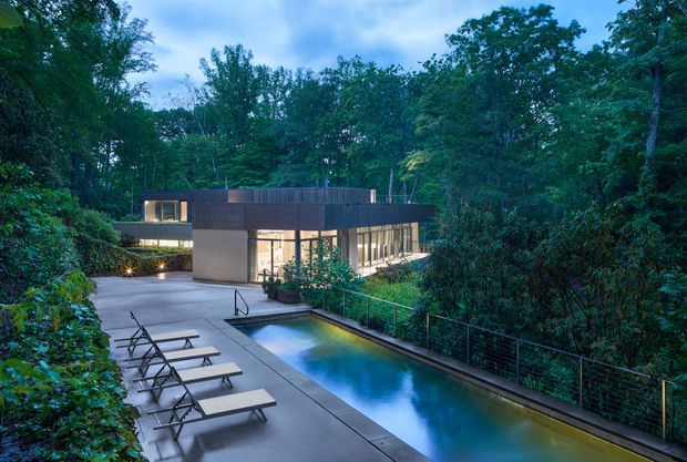 A modern, low-profile house nestled in dense woodland, viewed from an elevated outdoor terrace. The scene features a long narrow swimming pool, concrete paving, several reclining loungers, dark metal safety railing, and extensive glazing revealing the warmly lit interior. The building has a flat roof and simple rectilinear massing, creating a secluded contemporary retreat surrounded by mature trees.