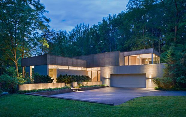 A sophisticated contemporary multi-level residence set within a wooded landscape, photographed at dusk. The home features clean rectilinear volumes, dark metal or panelled cladding, extensive warm-lit glazing, a recessed entry, landscaped retaining walls, and an integrated double garage with a broad paved driveway.
