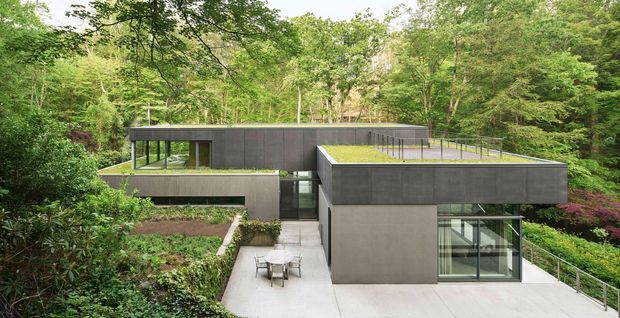 A modern, low-profile flat-roof residence nestled into a densely wooded landscape. The building is composed of crisp rectangular volumes with dark grey rendered or concrete-like walls, extensive floor-to-ceiling glazing, planted green roofs, and a pale concrete terrace and walkway. A small outdoor dining setting sits on the central paved courtyard, while mature trees and layered planting surround the home.