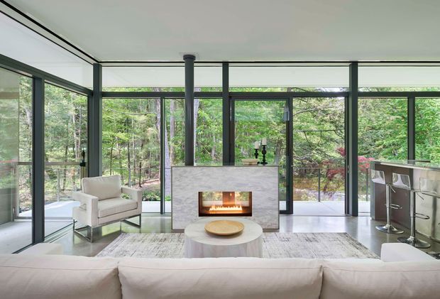 A bright, open-plan contemporary living room with floor-to-ceiling glazing overlooking a wooded setting. The room features a central pale stone fireplace with a lit fire, a light upholstered sofa and armchair, a round pale coffee table, a large neutral rug, dark slim-framed glazing, and a compact kitchen or bar area with stools to the right.