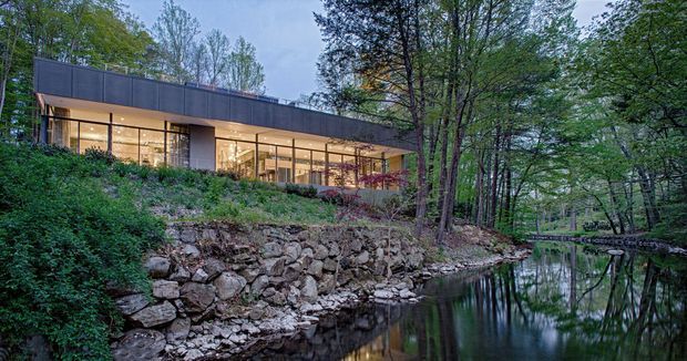 A contemporary single-storey pavilion-like house set into a wooded, sloping site beside a narrow reflective stream or pond. The building has extensive floor-to-ceiling glazing, a strong horizontal flat roofline, dark solid cladding or fascia, and warm illuminated interiors visible through the windows. A rough natural stone retaining bank and dense mature woodland surround the structure.