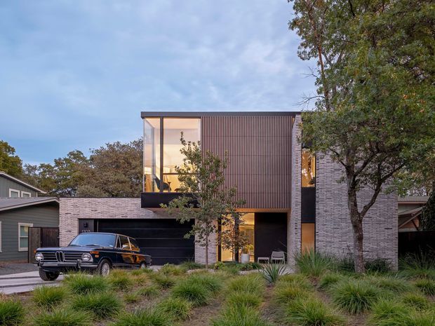 Exterior view of a contemporary multi-level residence with a rectilinear form, dark vertically ribbed cladding, pale brick walls, extensive glazing, and a recessed garage. The house is set within landscaped planting and photographed from the street, with a vintage car in the foreground.