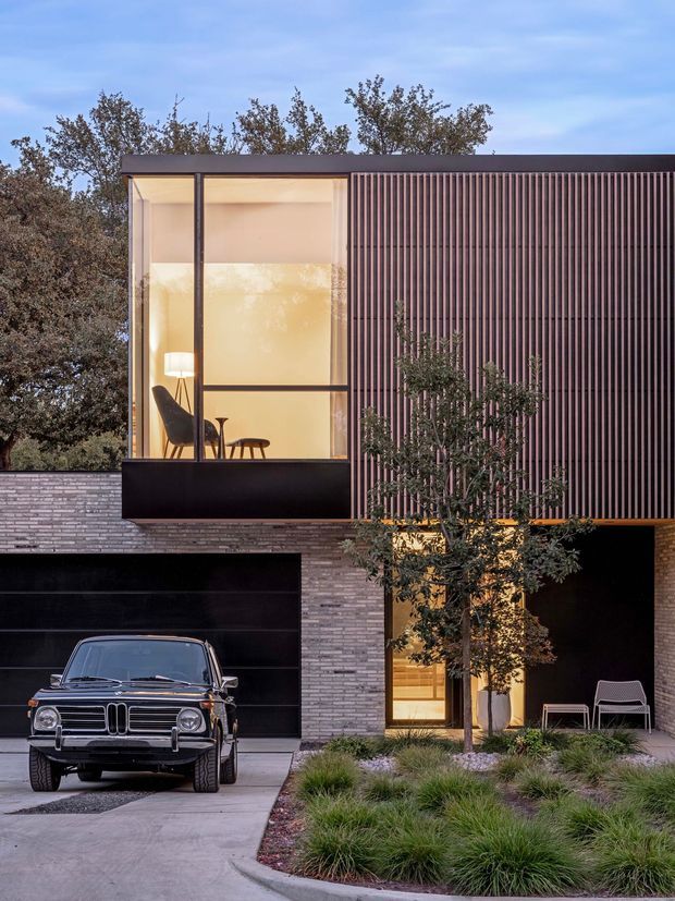 Photograph of a contemporary multi-storey residential facade at dusk, with a dark brick base, expansive black-framed upper-level glazing, and a prominent vertical timber or metal screening facade. Warm interior lighting reveals a minimalist sitting area behind the window. The house includes a large dark garage door, recessed entry, landscaped front garden, driveway, and a vintage car in the foreground.