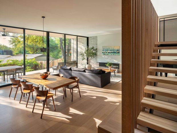 Bright, open-plan contemporary living and dining area with extensive floor-to-ceiling glazing overlooking a leafy suburban setting. A timber dining table with mid-century-inspired chairs sits beside a dark upholstered sofa, while a timber-framed open staircase and vertical timber screen define the right side of the room. Warm timber flooring, natural light, and restrained furnishings create an airy, refined interior.