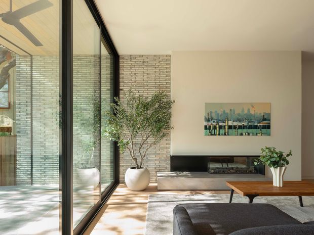 Bright contemporary living room with floor-to-ceiling black-framed glazing, a pale exposed-brick feature wall, built-in fireplace and low timber media cabinet. A dark sectional sofa, timber coffee table, indoor trees and cityscape artwork create a warm, refined interior with strong connection to the outdoors.