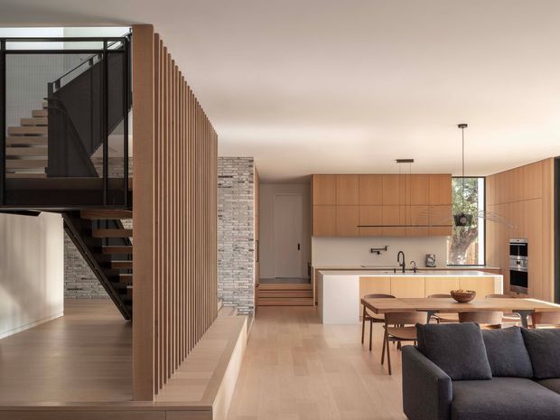 Open-plan contemporary interior featuring a timber-slatted screen partition, floating staircase with black metal balustrade, pale timber flooring, exposed light brickwork, and a warm wood kitchen with a central island. A dining table and dark sofa occupy the foreground, with abundant natural light from tall glazing.