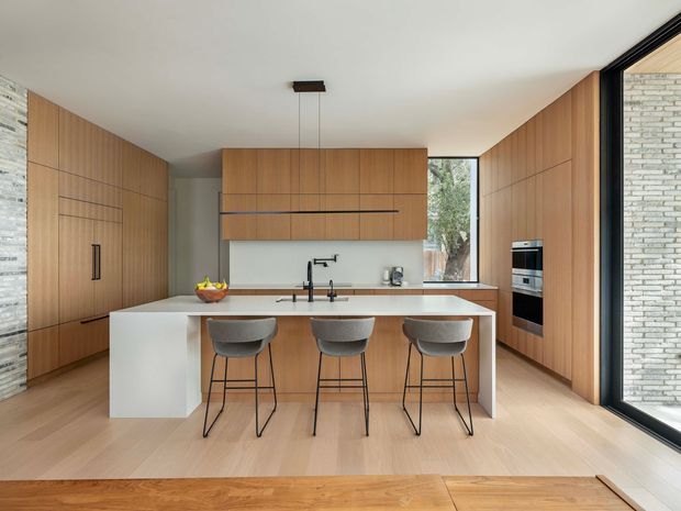 Contemporary kitchen featuring a large white island with seating for three, warm vertical timber cabinetry, integrated appliances, a pale stone-look benchtop, black tapware and window frames, and expansive glazing. The room has a clean, highly tailored open-plan appearance with timber flooring and a light neutral palette.