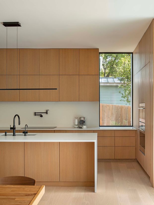 A warm, minimalist contemporary kitchen with extensive vertical-grain timber cabinetry, a pale stone island benchtop, integrated appliances, black plumbing fixtures, and light timber flooring. A large window brings in natural light and overlooks a fenced outdoor area. The kitchen includes a partial dining table in the foreground and streamlined pendant lighting.