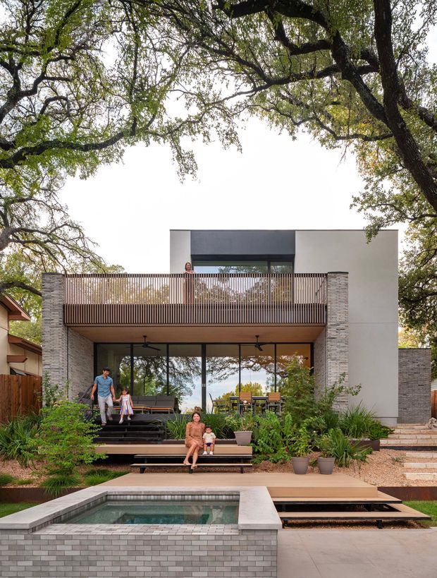 Contemporary two-storey residential house viewed from the landscaped rear garden, with a broad timber-slatted upper balcony, large glazed living area, pale rendered and masonry walls, stepped timber decking, lush planting, and a rectangular plunge pool in the foreground. Mature trees frame the property.
