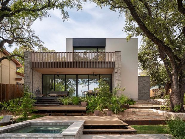 A contemporary multi-level residential home viewed from the landscaped rear garden. The façade combines pale rendered walls, dark metal-framed glazing, stacked natural stone-clad vertical elements, and a warm timber-lined balcony with slender vertical railings. Broad glazed doors open toward tiered timber and stone terraces, planted garden beds, and a small raised rectangular pool or spa in the foreground. Mature canopy trees frame the scene.