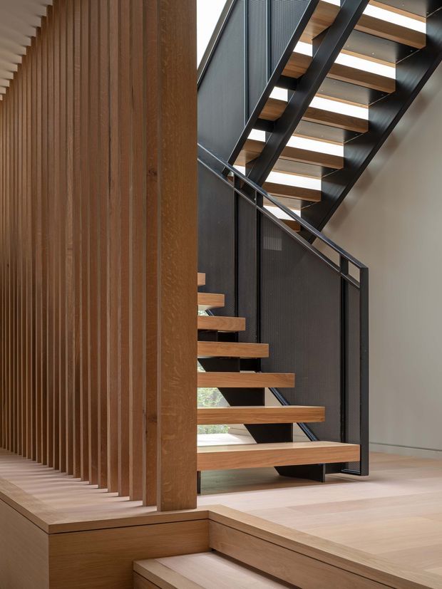 Interior architectural photograph focusing on a contemporary open-riser staircase. Warm timber treads and vertical timber slats contrast with a black metal stair structure, balustrade, and overhead framing. Pale timber flooring and a restrained neutral wall create a clean, refined, natural-toned composition.