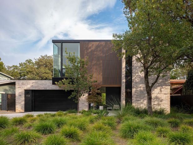 Contemporary multi-level residential facade with pale brick walls, a dark garage door, expansive glazing, and a prominent vertical timber screen. The house is set behind landscaped ornamental grasses and mature trees, with a flat or very low-pitched roofline and strong rectilinear volumes.