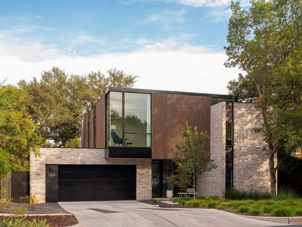 Contemporary two-storey residential facade set among mature trees, combining pale textured brick masonry, dark vertical timber screening, and expansive black-framed glazing. A wide dark garage door and concrete driveway occupy the lower level, while a projecting upper volume creates a strong geometric composition.